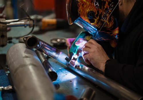 Worker in helmet with flames welding the steel part of a motorbike using an argon welding machine and gloves in a garage